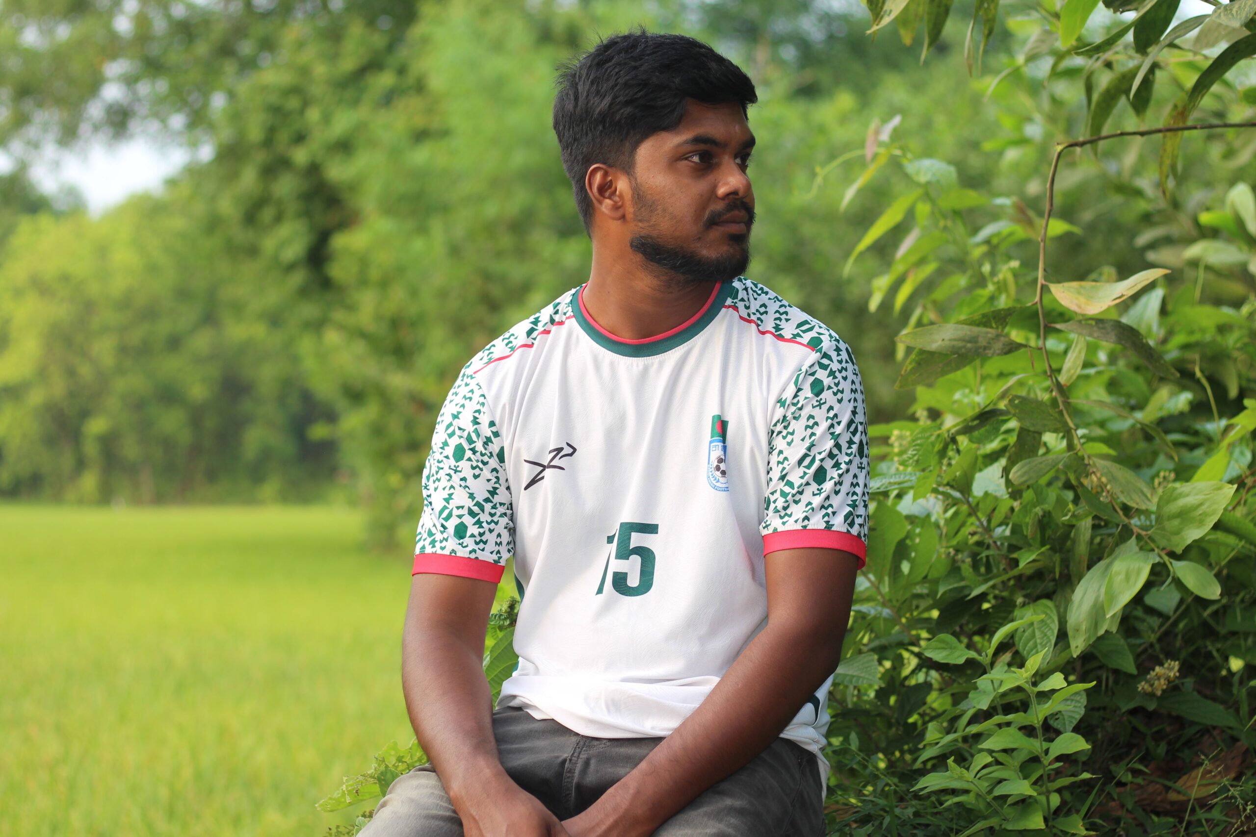 A young man in a sports jersey sitting amidst green foliage, enjoying a tranquil day.